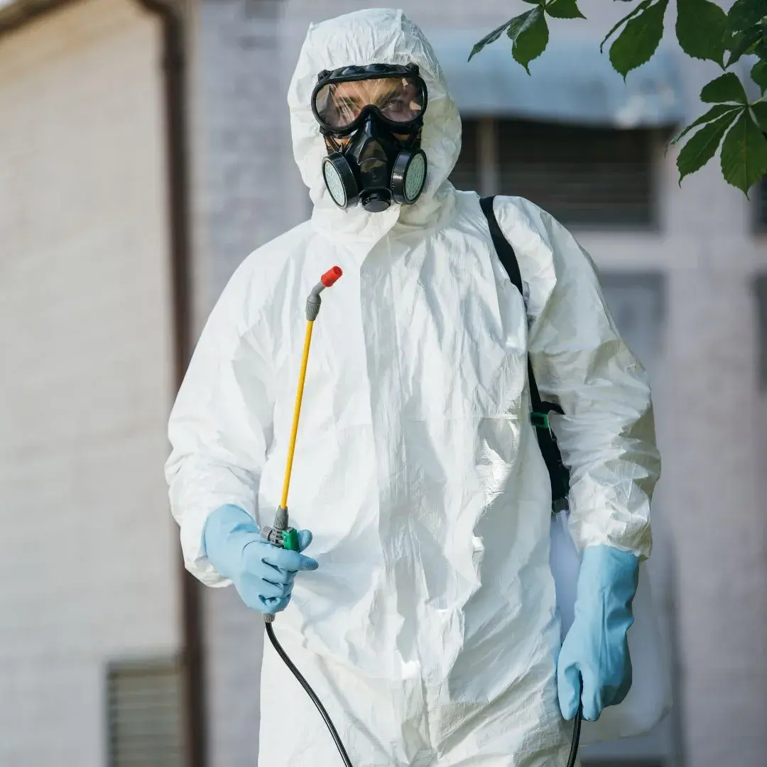 Person in protective suit and mask holding spray tool for pest control or sanitation outdoors.